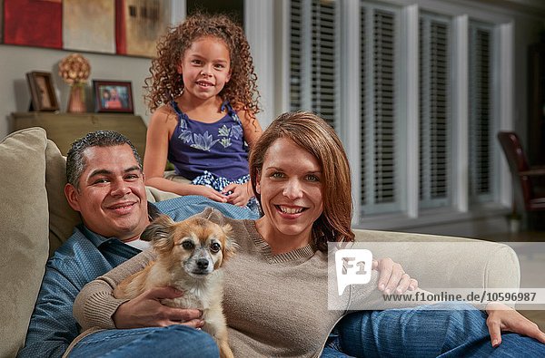 Parents and girl in living room on sofa with dog looking at camera smiling