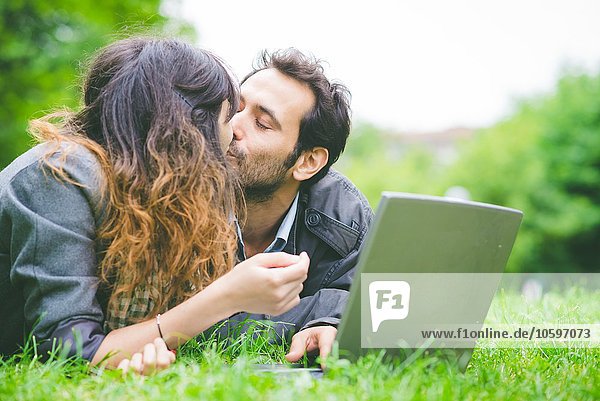 Head and shoulders of young couple lying on front on grass with laptop computer smiling