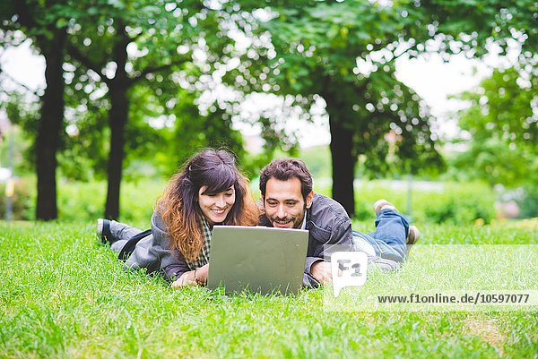 Front view of young couple lying on front on grass using laptop computer smiling