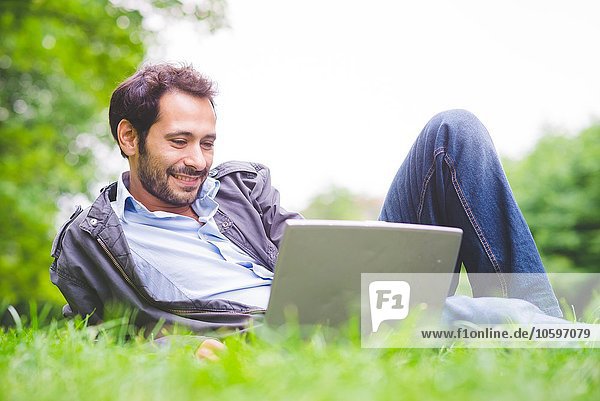 Junger Mann  der auf Gras liegt und lächelnd auf den Laptop schaut.