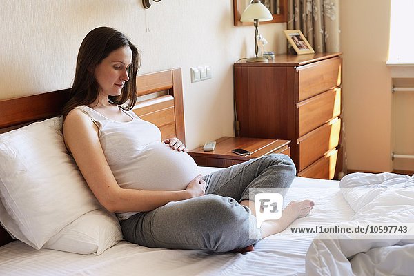 Schwangere Frau  die auf dem Bett sitzt und den Bauch hält.