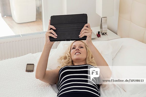 Hochwinkelansicht einer reifen Frau  die auf dem Rücken auf dem Bett liegt  mit erhobenen Armen und digitalem Tablett.