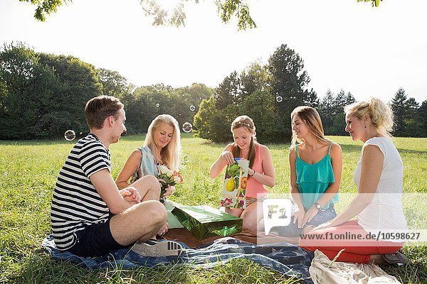 Family sitting on picnic blanket in park giving mature woman flowers and gifts smiling