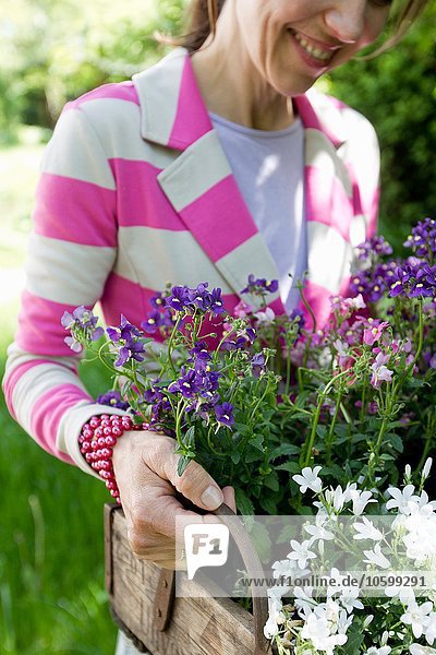 Reife Frau mit gestreifter Jacke  die eine Holzkiste mit lächelnden Blumen trägt.