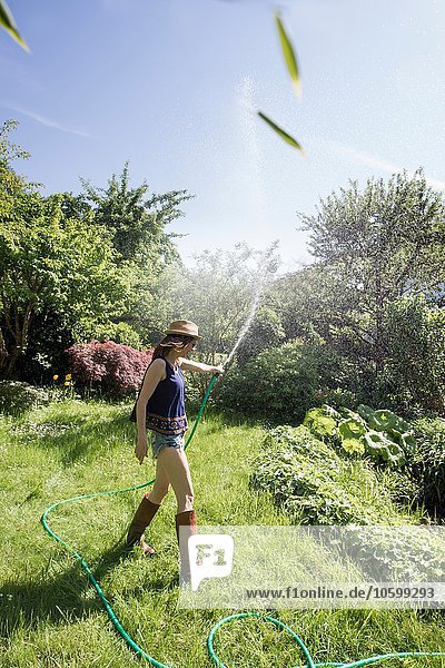 Seitenansicht einer reifen Frau im Garten  die mit einem Schlauch Wasser in die Luft spritzt.