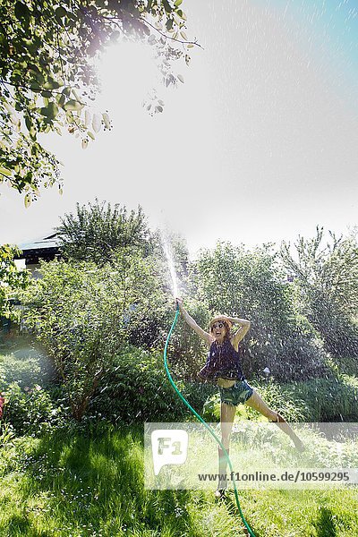 Vorderansicht einer reifen Frau im Garten  die auf einem Bein steht und mit einem Schlauch Wasser in die Luft spritzt.