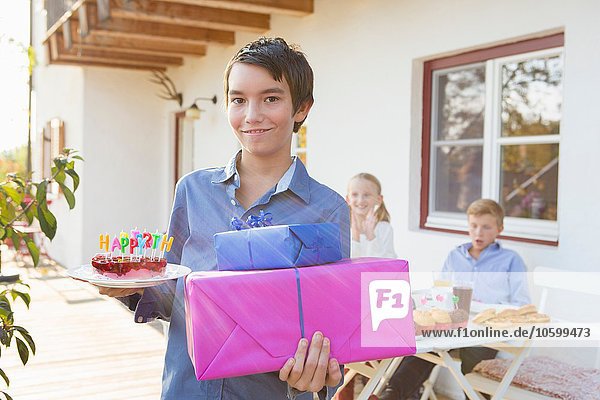 Portrait of teenage boy carrying birthday cake and birthday gifts on patio