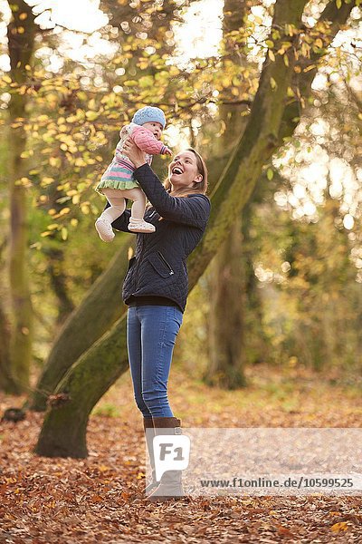 Mid adult woman holding up baby daughter in autumn park