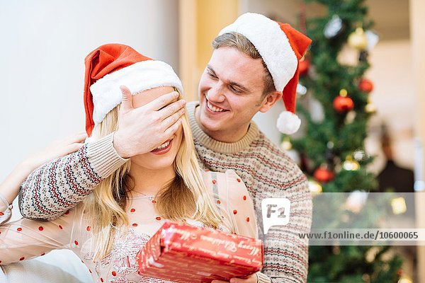 Junger Mann bedeckt die Augen der Freundin und überreicht ihr ein Weihnachtsgeschenk.