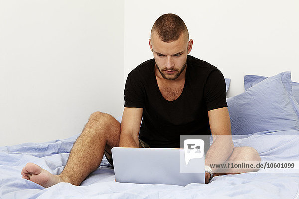 Mann sitzt auf dem Bett und benutzt einen Laptop