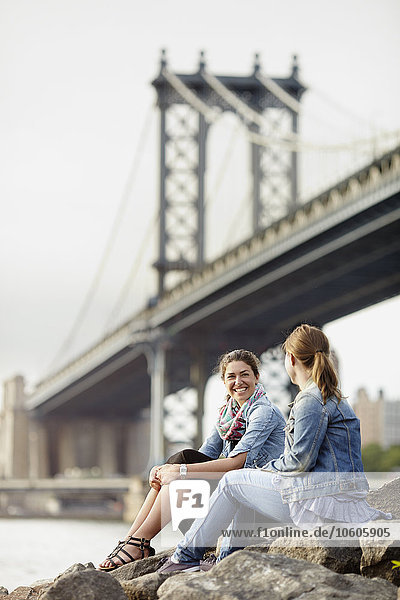 Zwei Touristinnen sitzen auf einem Felsen am Fluss mit der Brooklyn Bridge im Hintergrund