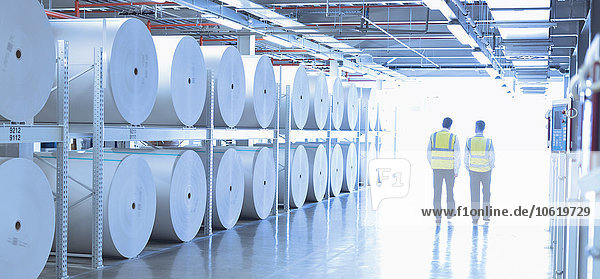 Arbeiter in reflektierender Kleidung  die in der Druckerei an großen Papierrollen entlanglaufen