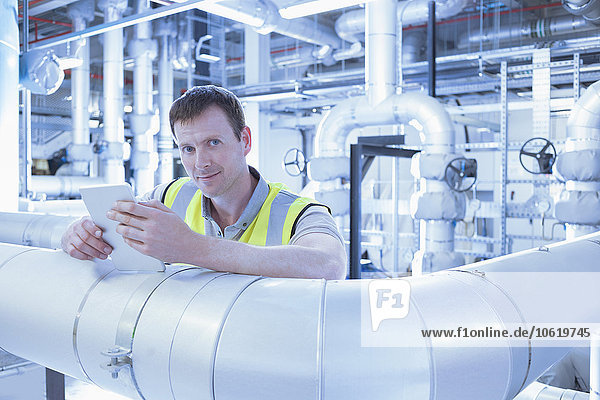 Porträt eines selbstbewussten Arbeiters mit einem digitalen Tablett  das sich in der Fabrik auf ein Rohr stützt.