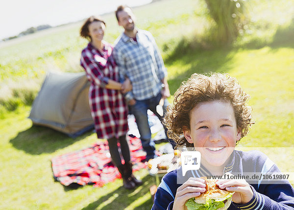 Portrait lächelnder Junge beim Hamburgeressen mit Eltern auf dem sonnigen Campingplatz