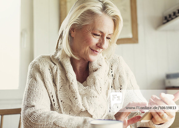 Lächelnde Seniorin im Pullover SMS mit Handy in der Küche