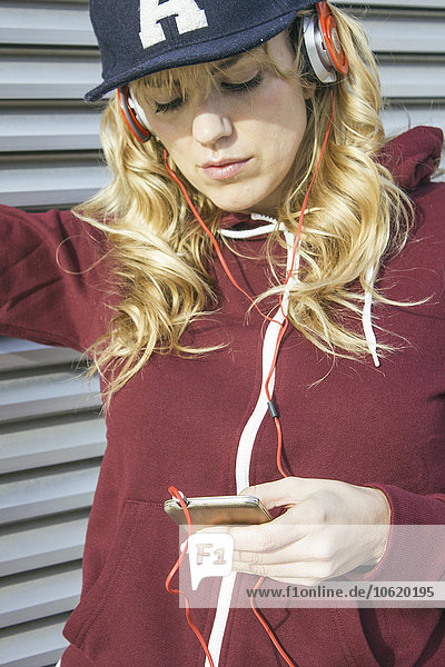 Blonde Frau mit Mütze und Kapuzenjacke beim Musikhören mit ihrem Smartphone