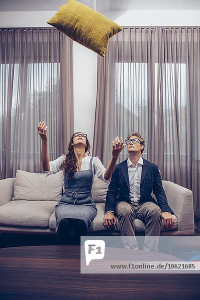Young couple at home sitting on sofa with cushion flying mid-air