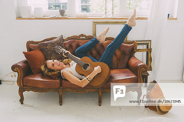 Happy woman lying on leather couch playing guitar