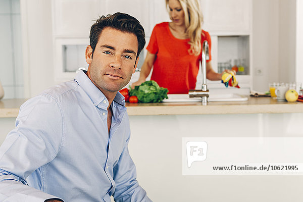 Porträt eines selbstbewussten Mannes mit Frau in der Küche im Hintergrund