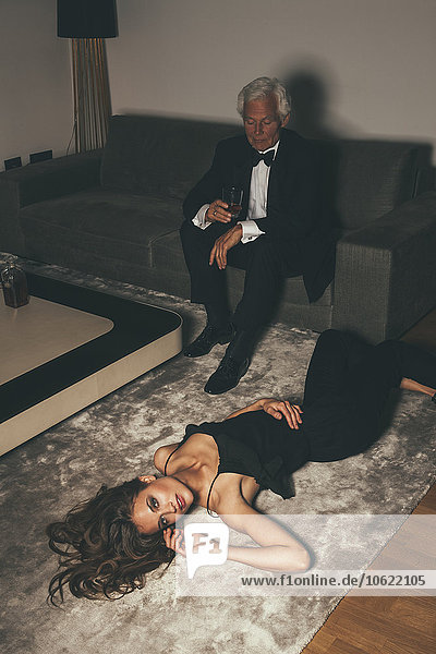 Senior man with drink on sofa looking at young woman lying on carpet