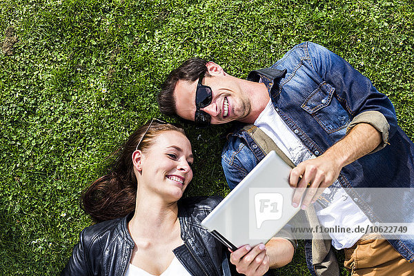 Glückliches junges Paar liegt auf der Wiese und schaut auf ein digitales Tablett.