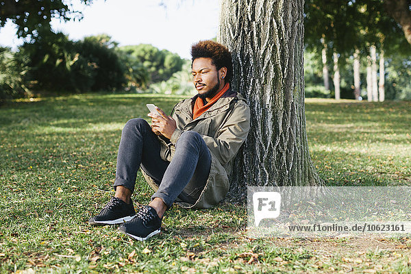 Junger Mann mit Smartphone an Baumstamm auf einer Wiese gelehnt