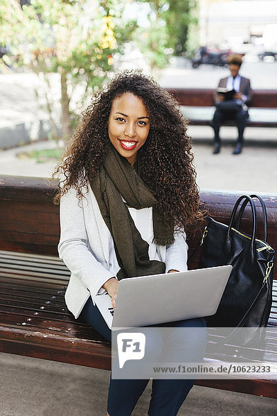 Lächelnde junge Frau mit Laptop auf der Bank