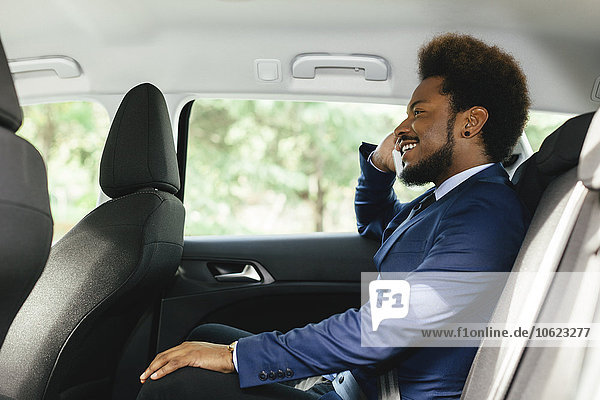 Lächelnder Geschäftsmann sitzt auf dem Rücksitz eines Autos und telefoniert mit dem Smartphone.