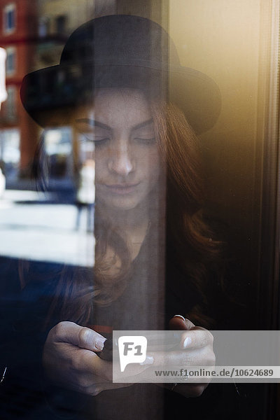 Junge Frau hinter der Fensterscheibe beim Blick aufs Handy