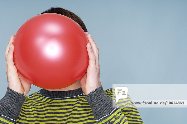 Teenager-Junge versteckt Gesicht hinter rotem Ballon vor blauem Hintergrund