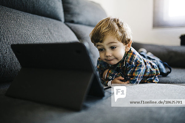 Porträt eines kleinen Jungen,  der auf der Couch liegt und auf ein digitales Tablett schaut.