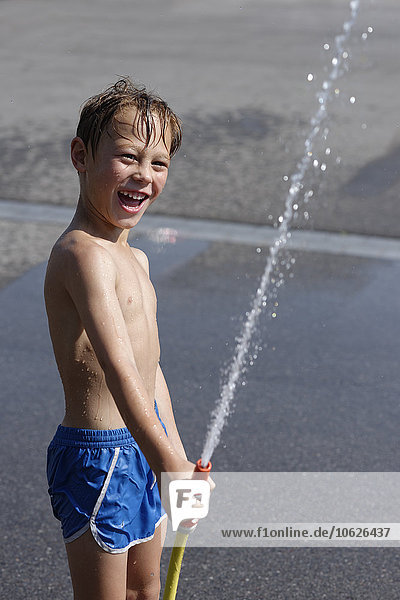Lachender Junge spritzt Wasser mit Gartenschlauch im Sommer