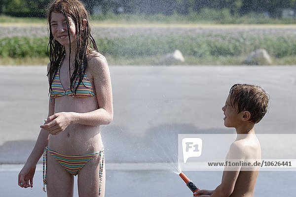 Kleiner Junge spritzt Wasser auf seine Schwester mit Gartenschlauch im Sommer