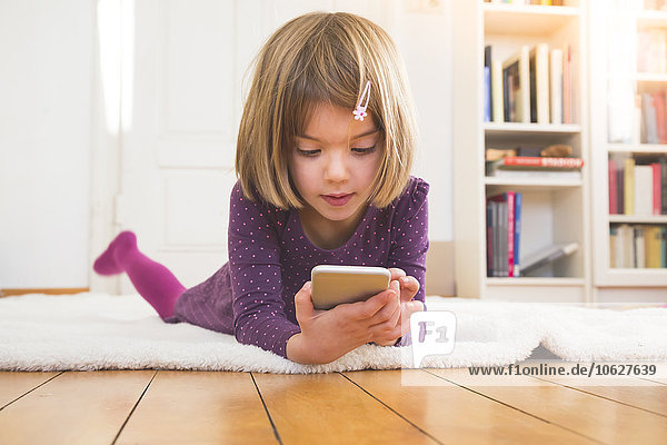 Kleines Mädchen liegt auf einer Decke auf dem Boden und schaut auf das Smartphone.