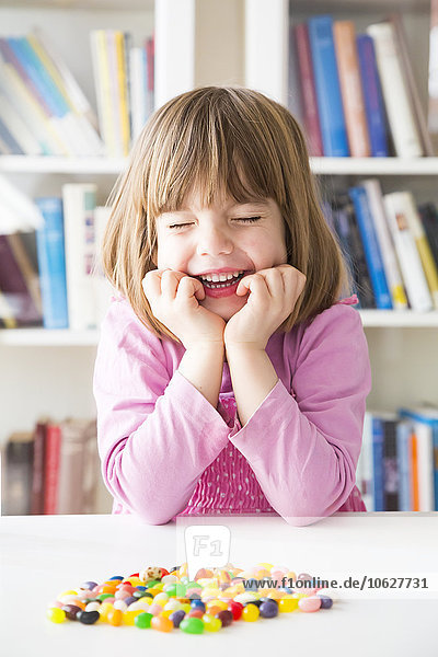 Porträt eines lächelnden kleinen Mädchens mit geschlossenen Augen und auf einem Tisch liegenden Geleebohnen.
