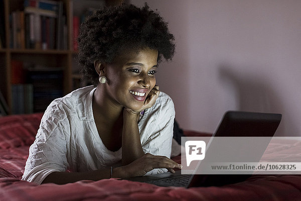 Porträt einer lächelnden jungen Frau auf dem Bett liegend mit Laptop
