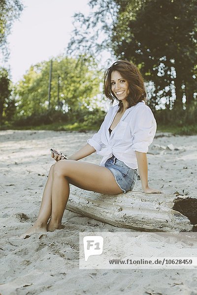 Porträt einer lächelnden Frau mit Smartphone auf Totholz am Strand