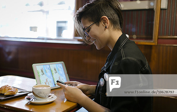 Junge Frau mit digitalem Tablett sitzt in einem Café und schaut auf ihr Smartphone.