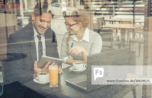 Zwei lächelnde Geschäftspartner sitzen in einem Coffee-Shop mit digitalem Tablett und Laptop