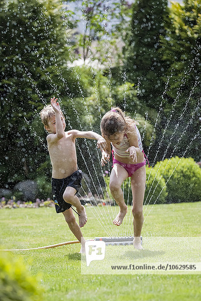 Children jumping over sprinkler in garden