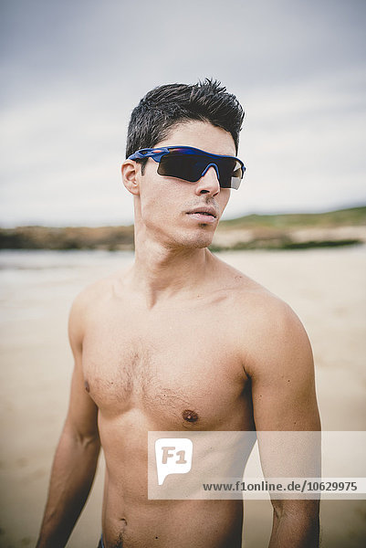 Porträt eines jungen Mannes ohne Hemd am Strand mit Sonnenbrille