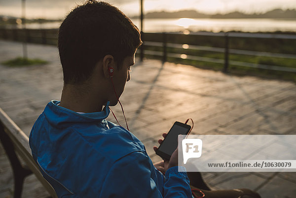 Athlet sitzt nach dem Training auf der Bank und hört bei Sonnenuntergang Musik vom Smartphone.