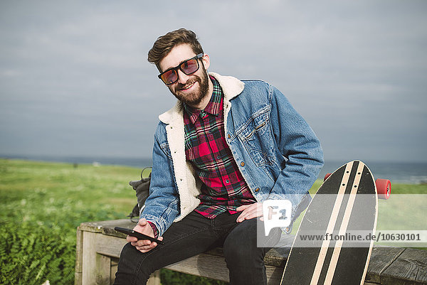 Junger lässiger Mann sitzt mit seinem Longboard im Park  lächelt und schaut in die Kamera.