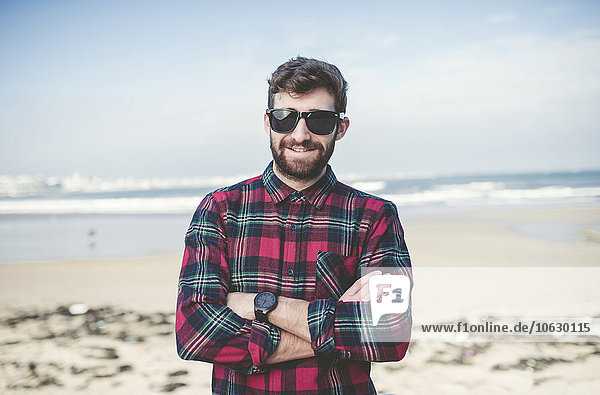Spain  La Coruna  portrait of smiling hipster with sunglasses standing on the beach