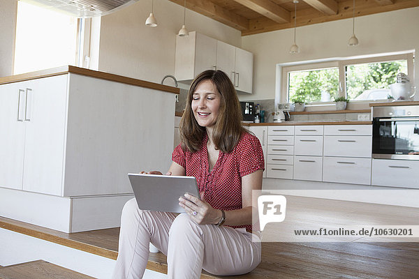 Reife Frau  die auf Küchenstufen sitzt  mit digitalem Tablett