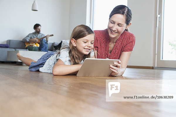 Mother and daughter lying on floor using digital tablet  father making music in background