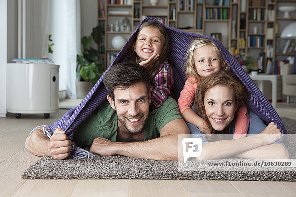 Porträt eines Paares mit ihren kleinen Töchtern  die zusammen auf dem Boden des Wohnzimmers liegen und von einer Decke bedeckt sind.