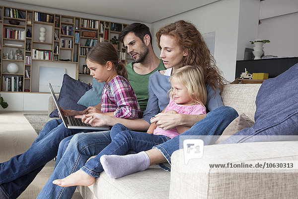 Paar sitzend mit ihren zwei kleinen Töchtern auf der Couch im Wohnzimmer mit Blick auf Laptop