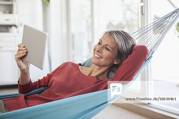 Lächelnde Frau zu Hause in der Hängematte liegend mit Blick auf digitales Tablett