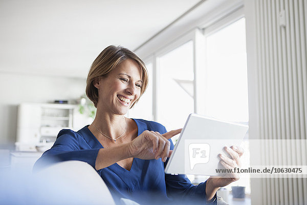 Lächelnde Frau zu Hause auf der Couch sitzend mit digitalem Tablett